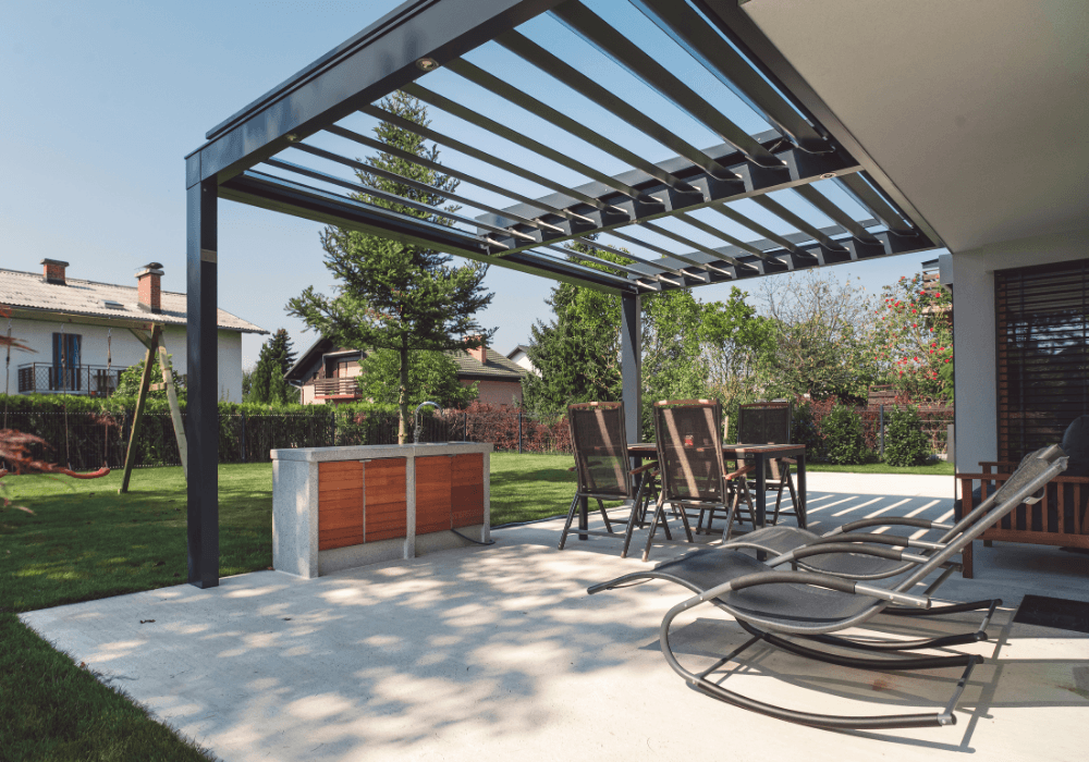 patio with a dark metal pergola, outdoor dining table and chairs, lounge chairs, and a built-in barbecue in a sunny backyard.