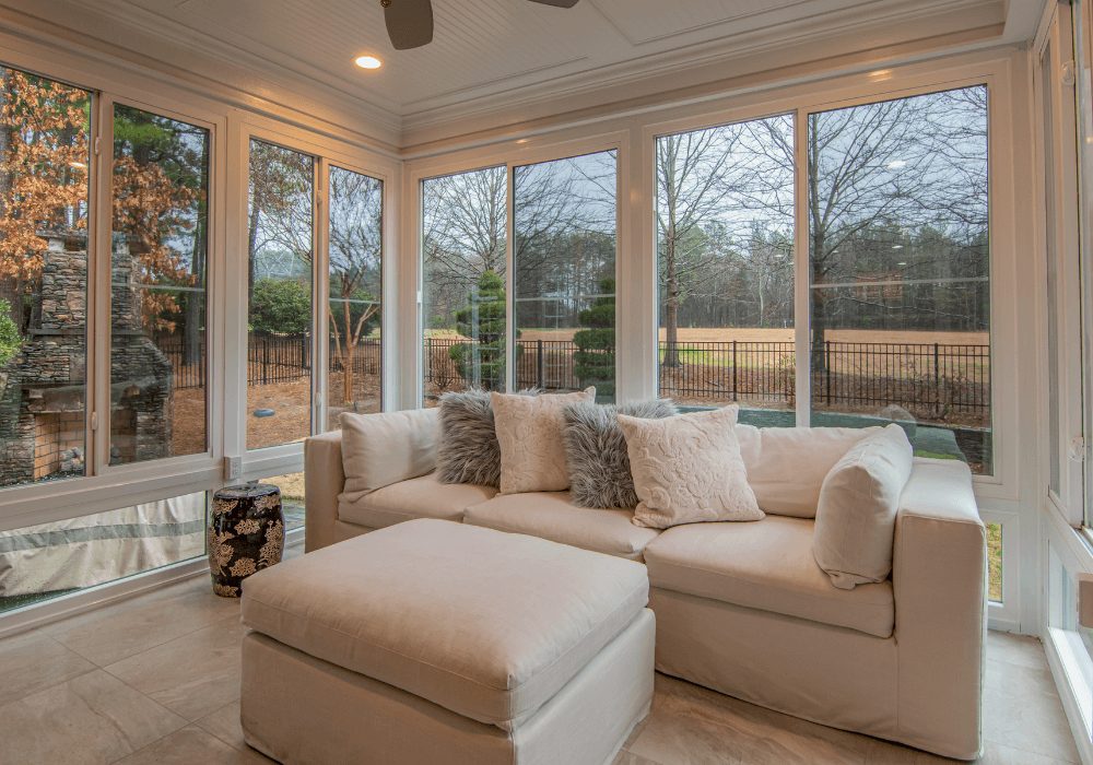 Bright sunroom with a light beige sectional sofa and matching ottoman, flanked by large windows showing a fenced yard and leafless trees outside, fluffy throw pillows add coziness.